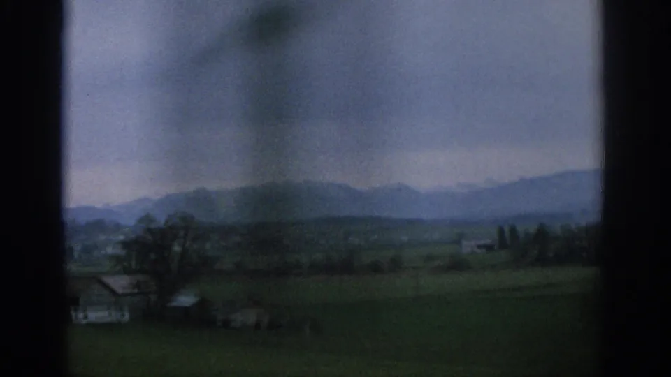 1963 Zurich-area rural landscape with farmhouses, green fields, bare trees, and distant mountains viewed through a window frame.