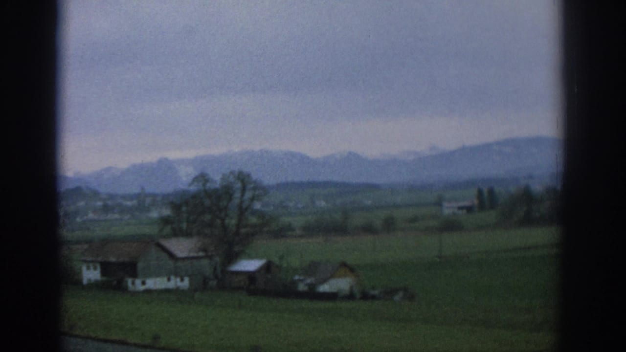 Farmhouses and green fields below a hazy mountain range, framed by a dark vertical window bar.