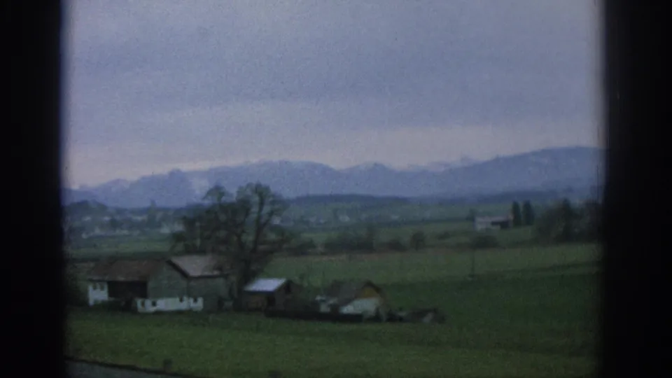 Zurich-area countryside in 1963 with low farm buildings, open fields, a bare tree, and hazy mountains beyond a window frame.