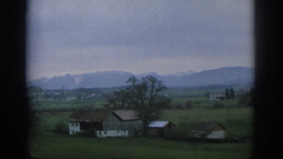 1963 Swiss rural view showing a farmhouse cluster in green fields, with bare trees and distant mountain ridges under cloud cover.