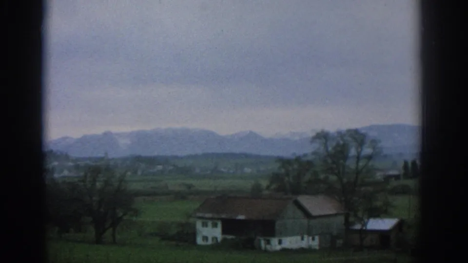 Zurich-area 8mm frame from 1963 showing farm buildings, winter-bare trees, open fields, and distant mountains under an overcast sky.