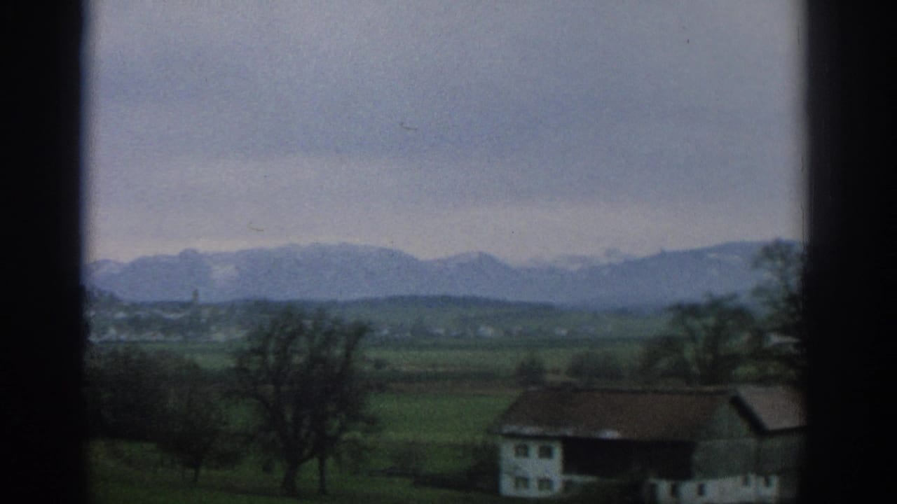 Window-framed view of green fields, a rural house, trees, and distant mountains.