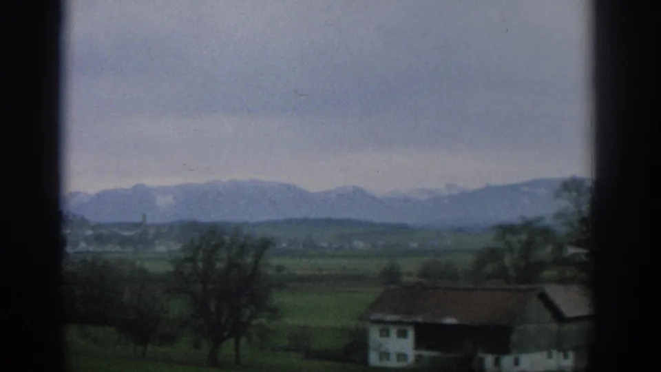 1963 Zurich-area home-movie frame showing Swiss countryside through dark window bars, with fields, a house, trees, and distant snow-topped mountains.