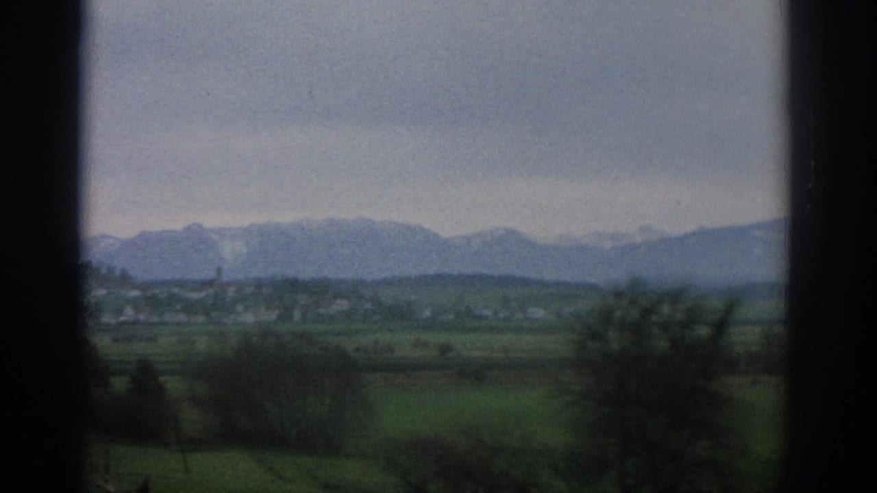 Countryside beyond window bars with trees, fields, village buildings, and mountains.