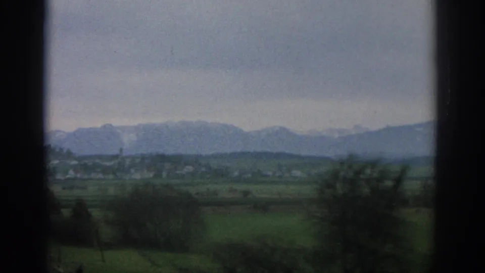 1963 Zurich-area countryside seen from behind window framing, with green fields, scattered trees, low buildings, and hazy mountains.