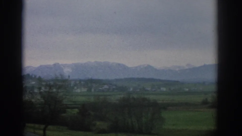 1963 Zurich-area 8mm frame with soft focus on open fields, bare trees, distant settlement, and a snow-dusted mountain range.