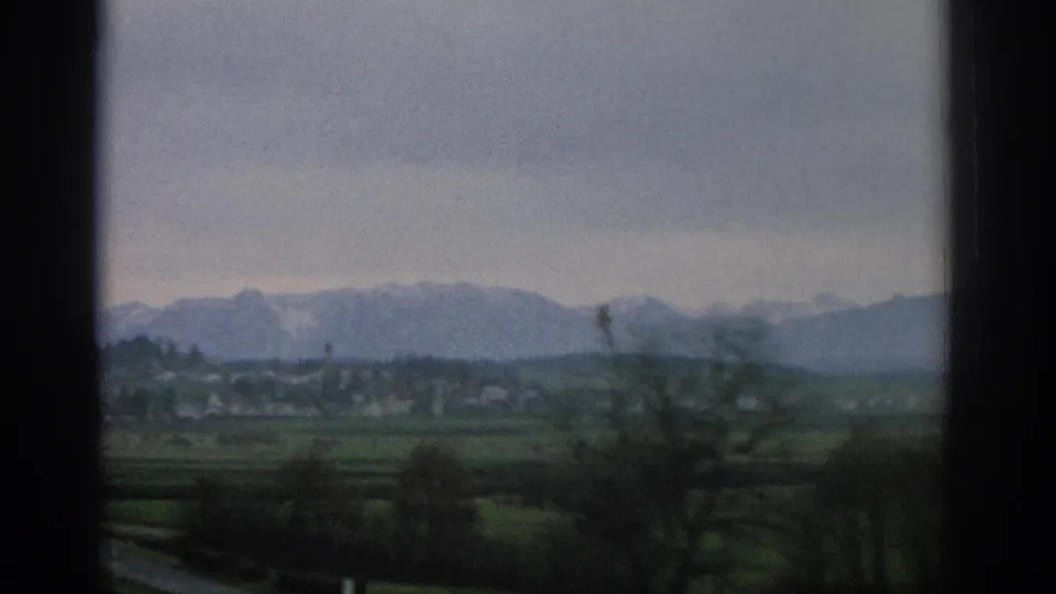 1963 Zurich-area home-movie frame looking through window bars across fields, trees, a distant village, and muted mountain peaks.