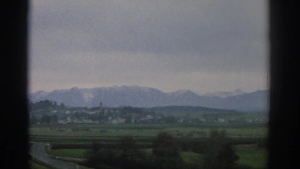 1963 Swiss countryside view with green fields, a small distant village, and snow-capped mountains seen through dark vertical borders.
