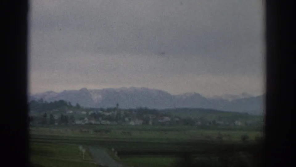 Soft 8mm view of green Swiss farmland and a distant settlement with snow-touched mountains beneath an overcast sky.