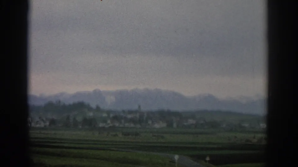 Cloudy 1963 Switzerland landscape with green fields, scattered valley buildings, and hazy alpine peaks in soft focus.