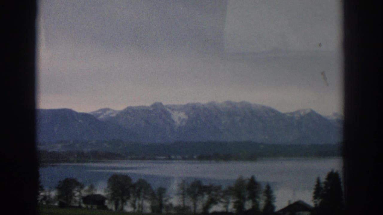 Window-framed lake view with distant mountains and trees along the shore.