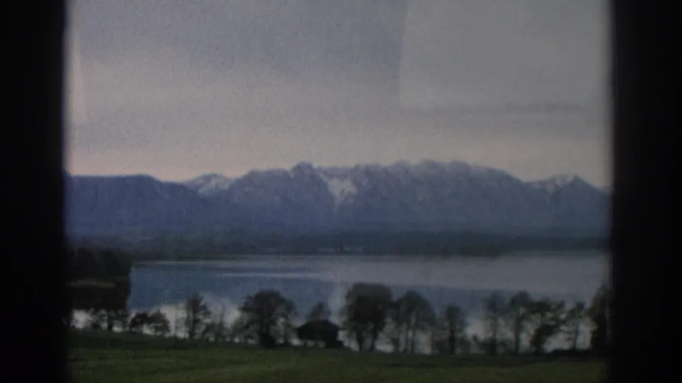 The 1963 home-movie frame shows grass, lakeside trees, still water, and a hazy alpine ridge beyond the window.