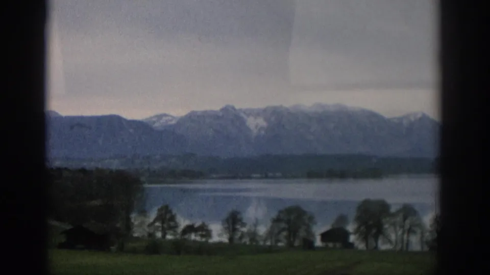 A window-framed 8mm landscape near Zurich with lakeside trees, small structures, water, and snow-dusted mountains.