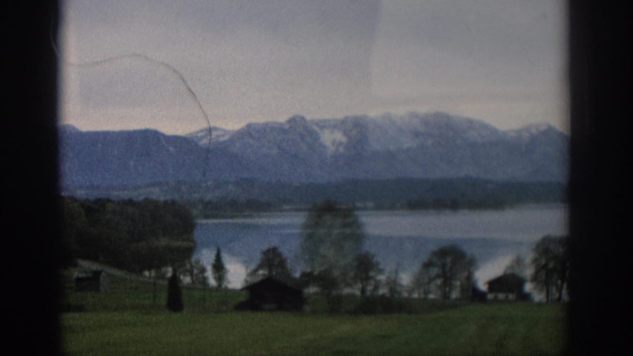1963 Zurich home-movie view of a calm lake, dark tree line, green foreground, and snow-capped mountains under an overcast sky.
