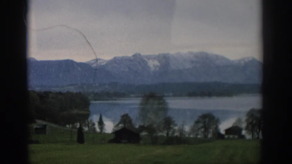 1963 Zurich home-movie view of a calm lake, dark tree line, green foreground, and snow-capped mountains under an overcast sky.