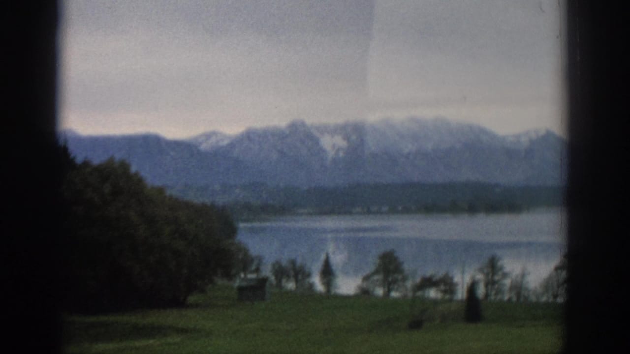 Lakeside field with trees and mountains beyond the water.