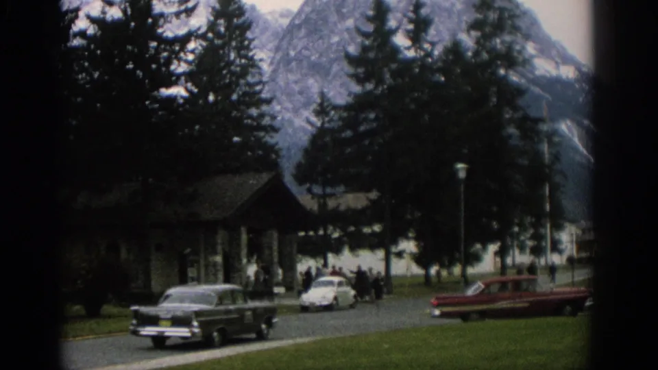 1963 Zurich roadside scene with classic cars, distant pedestrians, a small lakeside building, tall trees, water, and mountains behind.