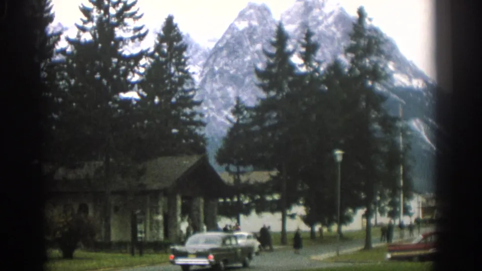 Zurich-area 8mm frame showing a classic car by a lakeside road, distant people, trees, a small structure, and steep mountain slopes.