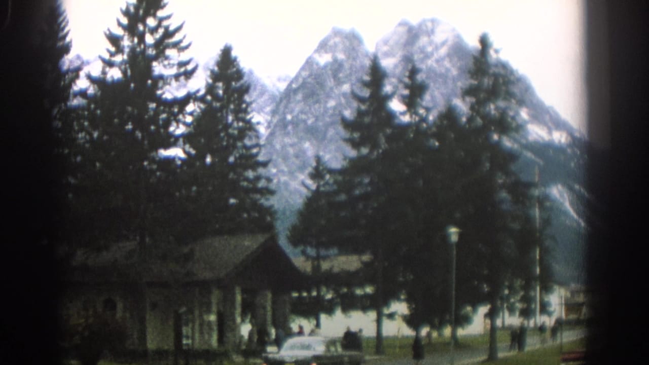 1963 Zurich park view with evergreens, distant snowy mountains, a low building, pedestrians, and road traffic in soft 8mm color.