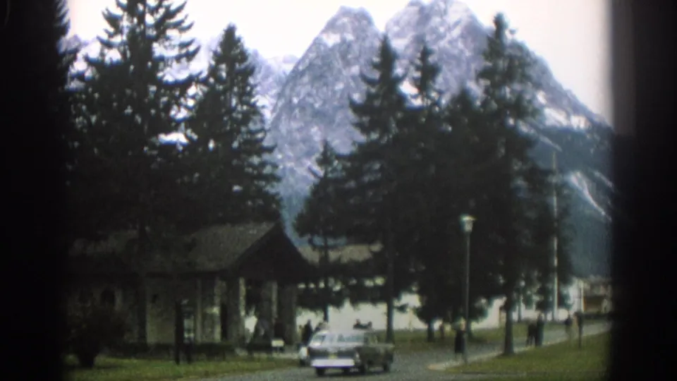 A white compact car and a dark sedan move through a Zurich park setting with pedestrians, evergreens, and mountains behind them.