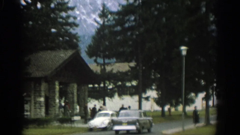 1963 Zurich footage shows two cars passing a stone park building while people stand near the road and mountains rise behind trees.