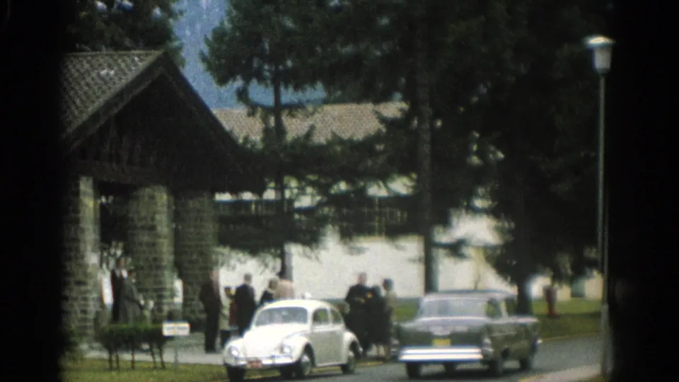 Near the end of the clip, two cars pass people gathered by a stone building in a Zurich park, with trees and mountains in the background.