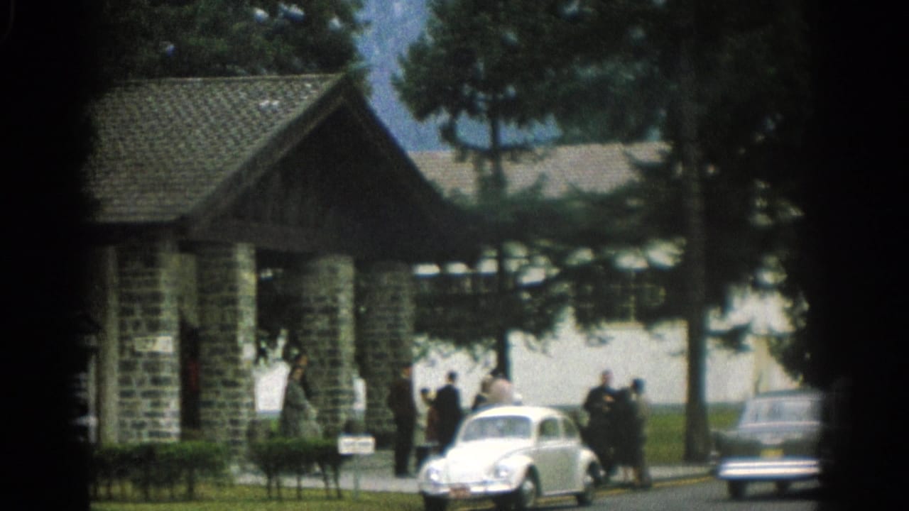 1963 Zurich home-movie frame showing a stone-and-timber entrance structure, trees, people, and parked mid-century cars.