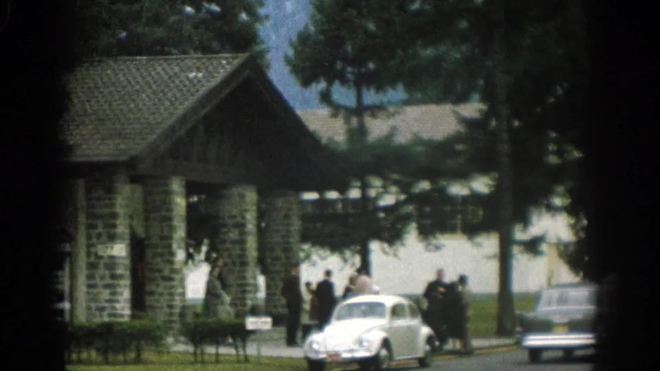 1963 Zurich home-movie frame showing a stone-and-timber entrance structure, trees, people, and parked mid-century cars.