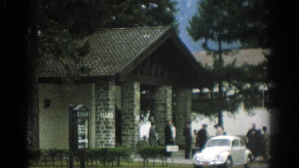 1963 Zurich frame with a signpost, lodge-like gate structure, people at the entrance, and a white vintage car.