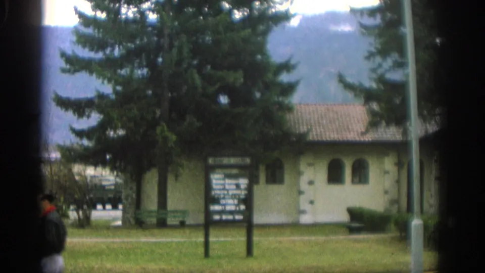 1963 Zurich home-movie frame showing a lawn sign, pale building, benches, trees, and mountain backdrop.