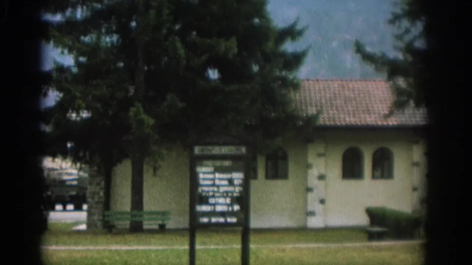 1963 Zurich frame centered on a signboard, benches, evergreen trees, and a pale building in a landscaped setting.