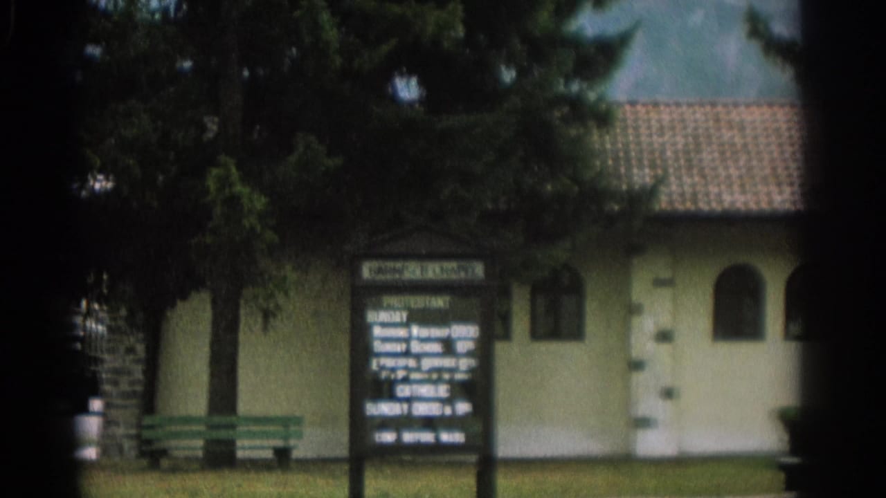 Small chapel exterior with service sign, trees, lawn, and green bench.