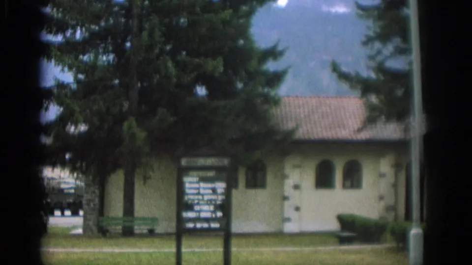 Wider 1963 Zurich home-movie view of the chapel grounds, with the sign at left, trees overhead, a green bench, and mountains behind the roofline.