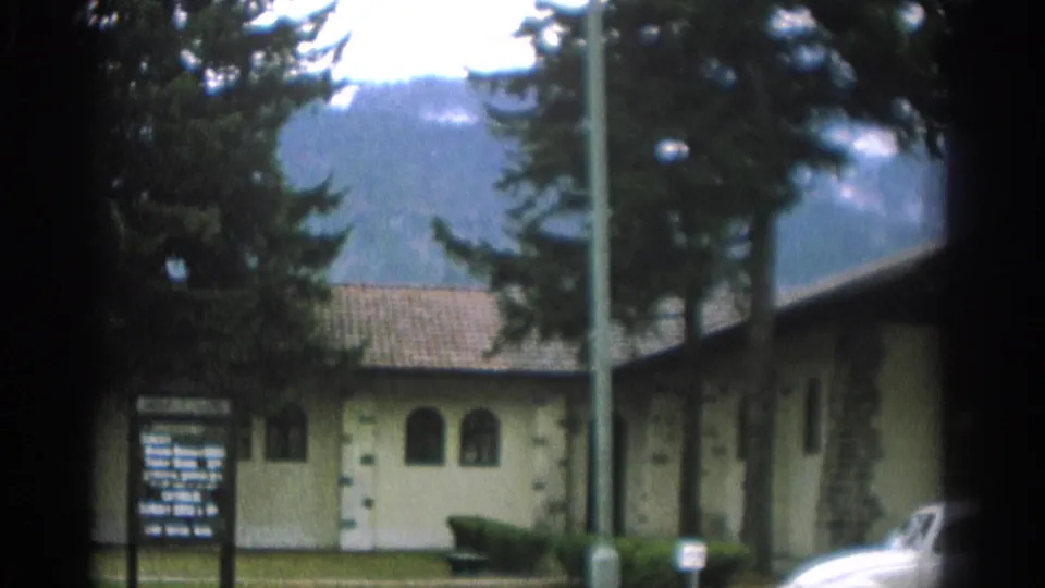 Late frame from the 1963 Zurich chapel clip, showing the service sign, chapel corner, trees, mountains, lawn, bench, and a partial car at lower right.