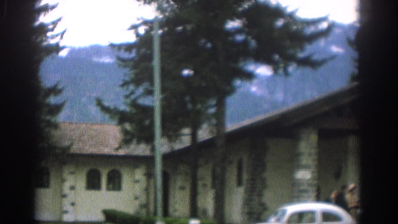 1963 Zurich home-movie frame showing a stone-and-stucco building, evergreen trees, mountain backdrop, and a partly visible white classic car.