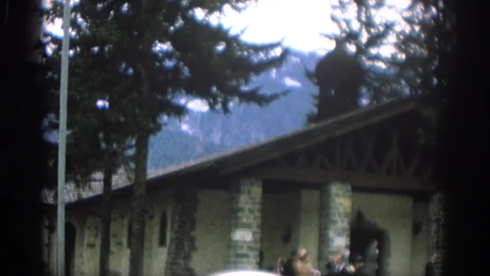 1963 Zurich frame with a building entrance, stone columns, evergreens, distant mountains, and a few people gathered outside.