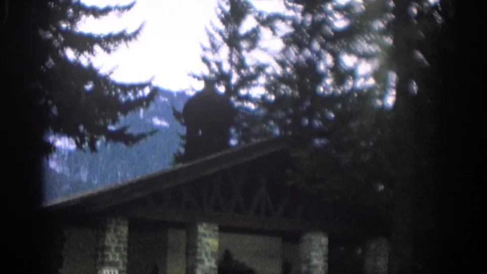 1963 Zurich frame showing a darkly framed view of the building roofline, stone columns, evergreen trees, mountains, and bright sky.