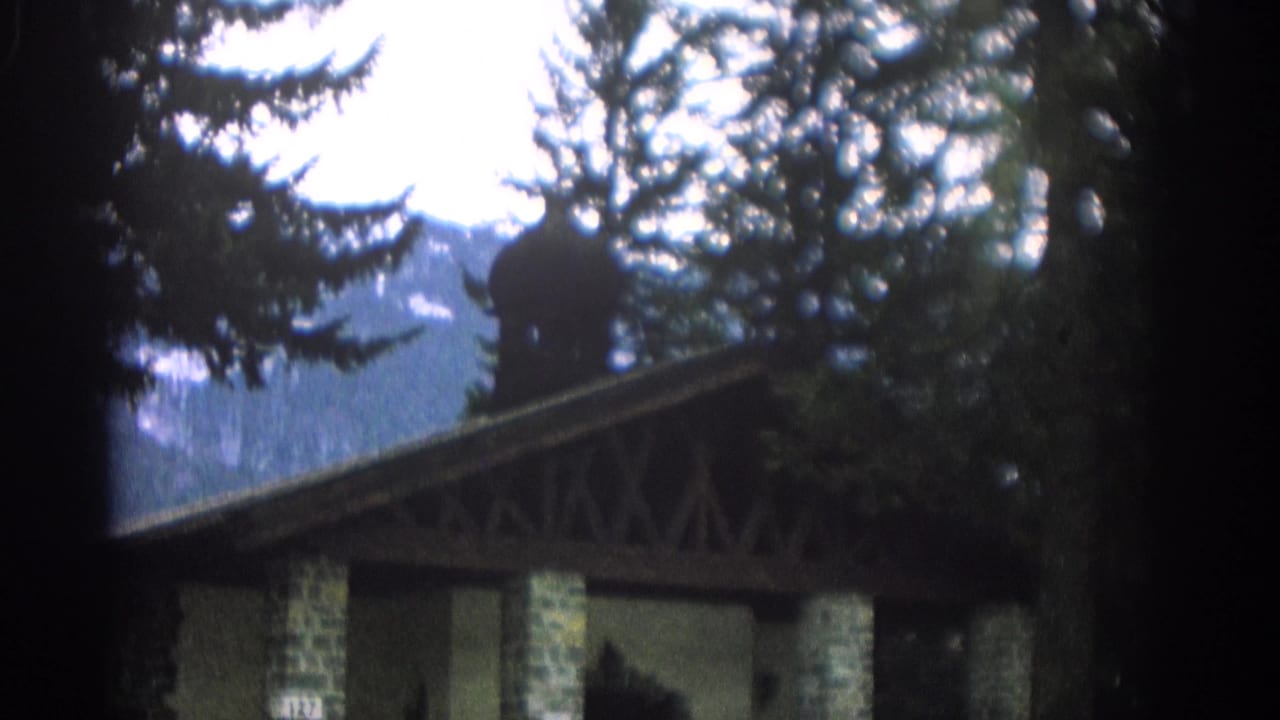 1963 Zurich home-movie frame of a stone-and-timber pavilion among evergreens with alpine mountains in the background.