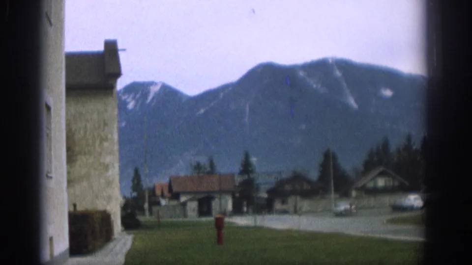 The camera turns from the pavilion toward an alpine view with nearby buildings, road, lawn, and parked cars in Zurich, Switzerland.