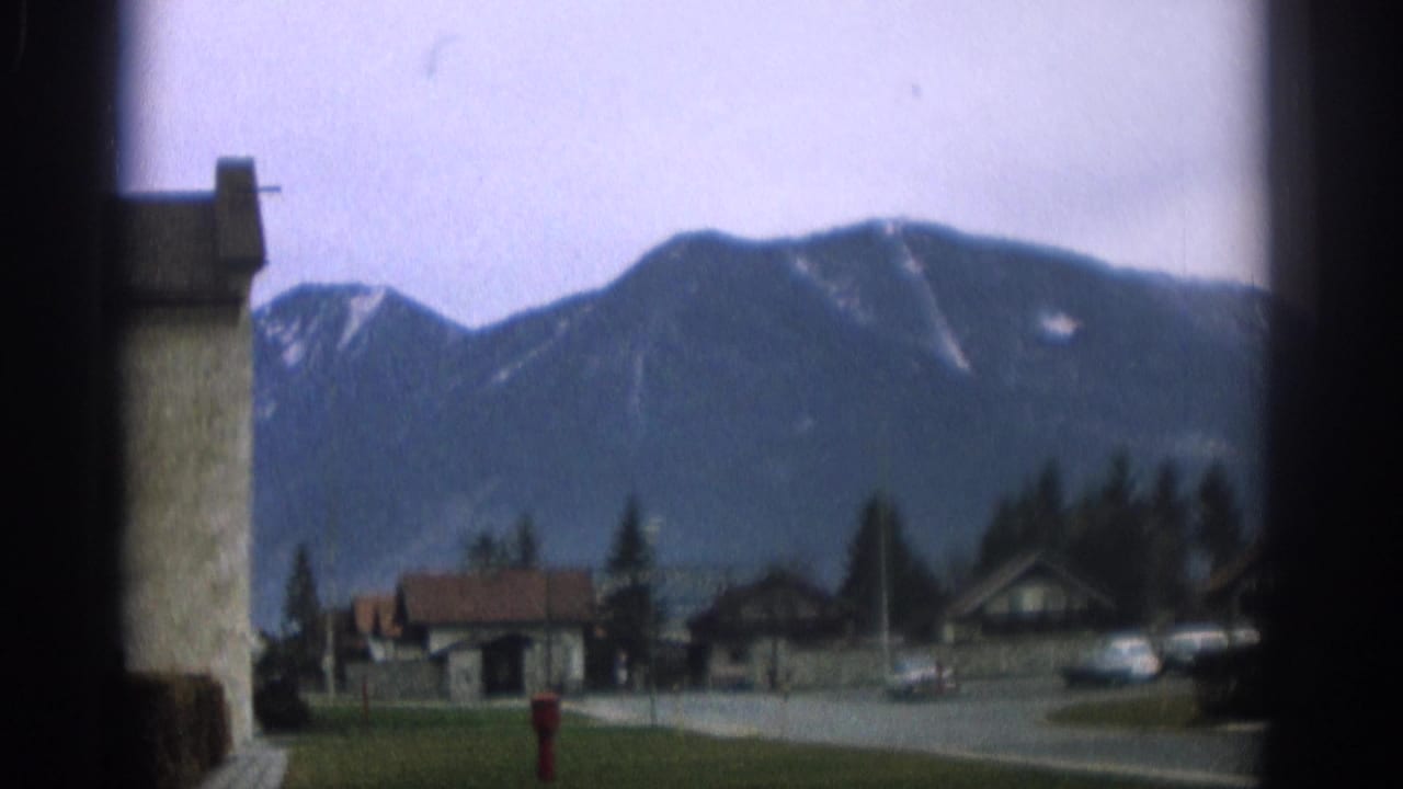 Mountain town street with houses, parked cars, trees, and snow-dusted peaks behind dark frame edges.
