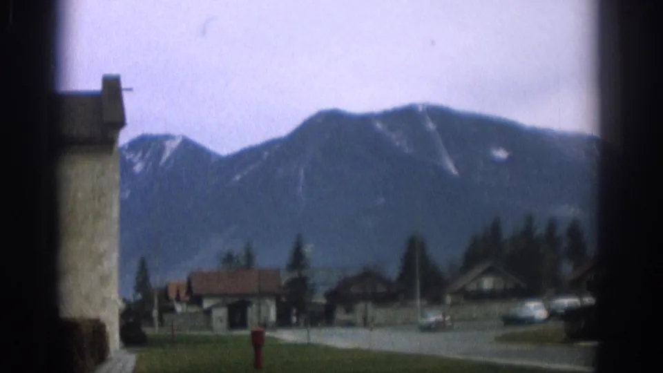 1963 Zurich-area home-movie view of a quiet mountain town street with houses, cars, trees, and snow-dusted peaks beyond.