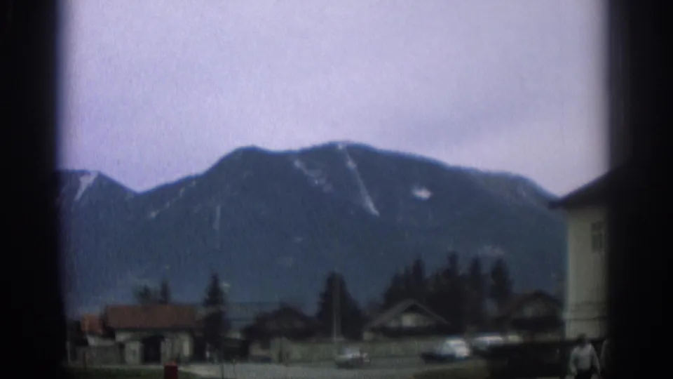 1963 Zurich-area town view with an alpine mountain backdrop, small buildings, trees, cars, and a dark window or gate edge.