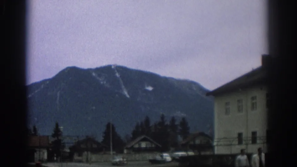 1963 Zurich-area home-movie frame showing a mountain town with chalet-like houses, trees, cars, and a pale building at right.