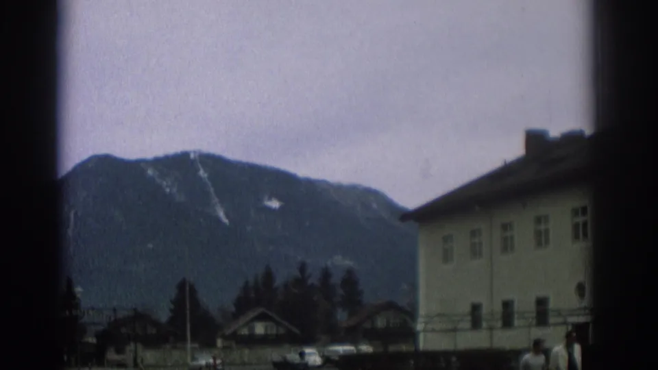 1963 Zurich-area street view with mountains, village buildings, parked cars, trees, and a few distant pedestrians near a pale building.