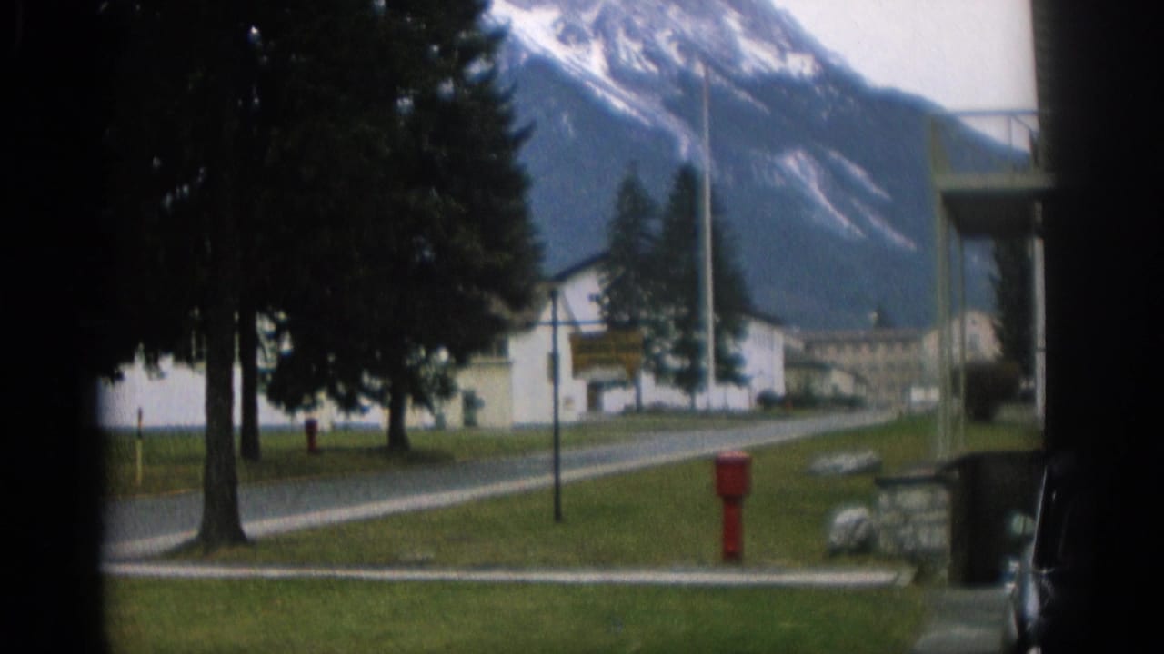Trees, a small white building, a paved road, a red hydrant, and snow-streaked mountains.