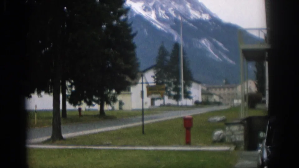 1963 Swiss 8mm frame showing evergreen trees, a small white roadside building, a red hydrant, and mountains beyond.