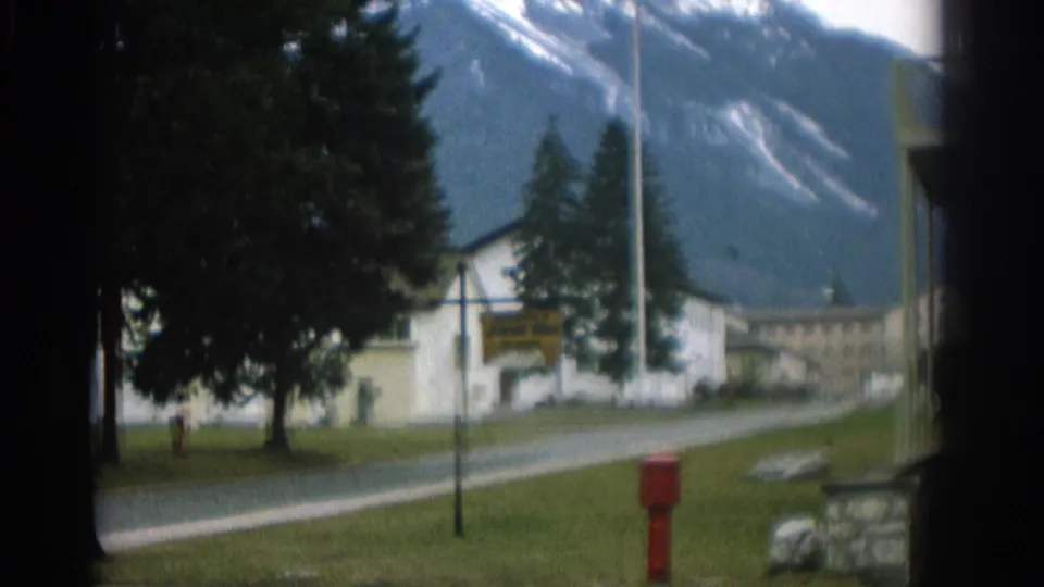 1963 Zurich-area home-movie frame with a white building, roadside sign, red hydrant, and mountain slope in the background.