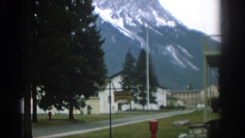 1963 Swiss roadside view with a curving road, green lawn, tall evergreens, white building, red hydrant, and mountains behind.