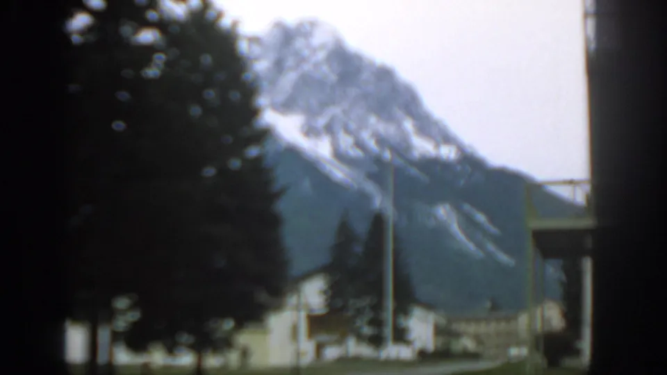 Soft 1963 Zurich-area frame showing blurred foreground trees, a white roadside building, a utility pole, and a snow-streaked mountain.