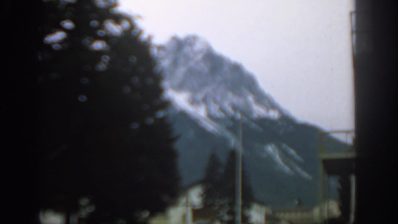 1963 Swiss 8mm frame showing a snow-streaked mountain partly obscured by dark evergreens, with a small structure below.